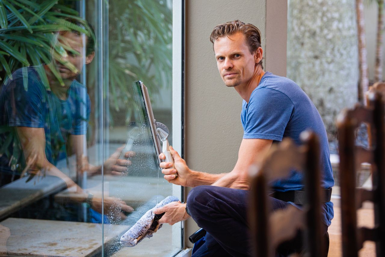 Un homme en t-shirt bleu nettoie une grande vitre à l’intérieur avec une raclette.